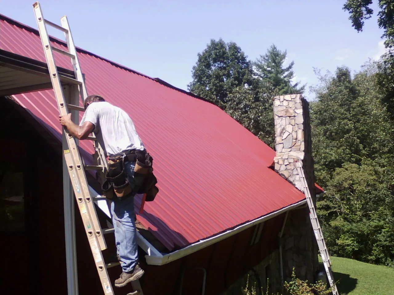 Roofer on a ladder beside a red metal roof and stone chimney — Soffit & Fascia Repair by Roofing Brothers in Morriston
